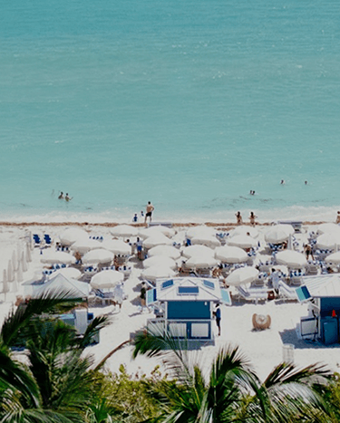 Panoramic view of a crowded beach with turquoise water, white sand, colorful umbrellas, and palm trees.